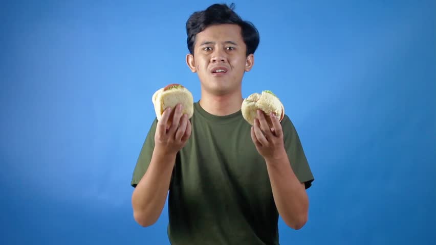 Asian man holding two hamburgers and choosing to eat them greedily, isolated on blue background