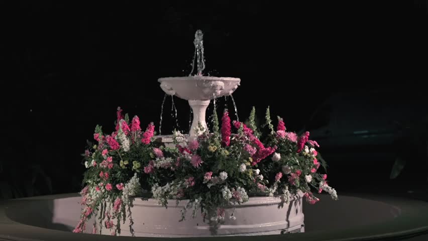 Ornate white tiered water fountain sprays upward, surrounded by a lush arrangement of pink, white, and yellow flowers at night for an outdoor event.