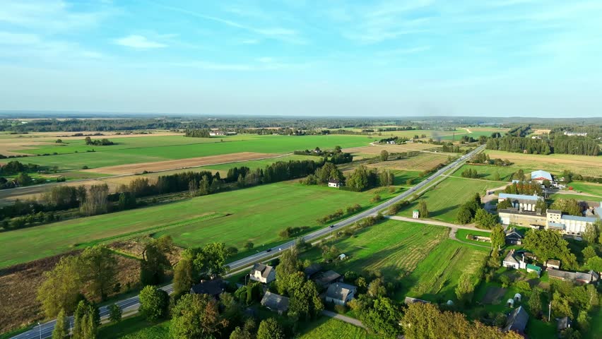 Aerial view of a long country road cutting through wide open farmland, green fields, and scattered rural homes under soft late-afternoon light.