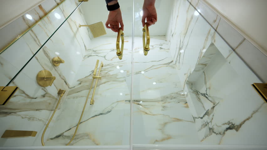 POV of opening the glass doors of the shower in the luxury bathroom with elegant gold fixtures. The marble-style wall tile adds a high-end, sophisticated look.