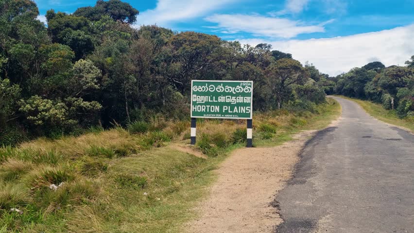 Entrance sign of Horton Plains National Park with an empty road leading inside, surrounded by highland vegetation under a shiny blue sky at midday, conveying calm nature, conservation