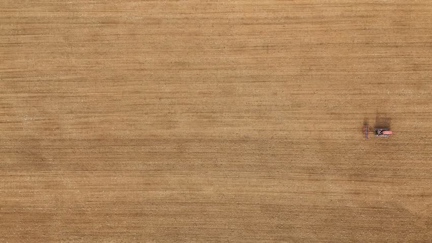 Top down shows a small crawled tractor working a large, golden‑brown field in rural Tuscany, Italy, smoothing and leveling the soil in gently rolling farmland bordered by a strip of green vegetation.