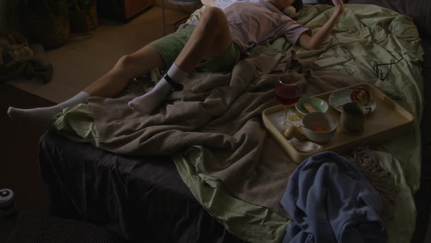 High angle shot of young woman, wearing house arrest ankle monitor, dancing to music, listening to music in large headphones and lying in bed with pizza