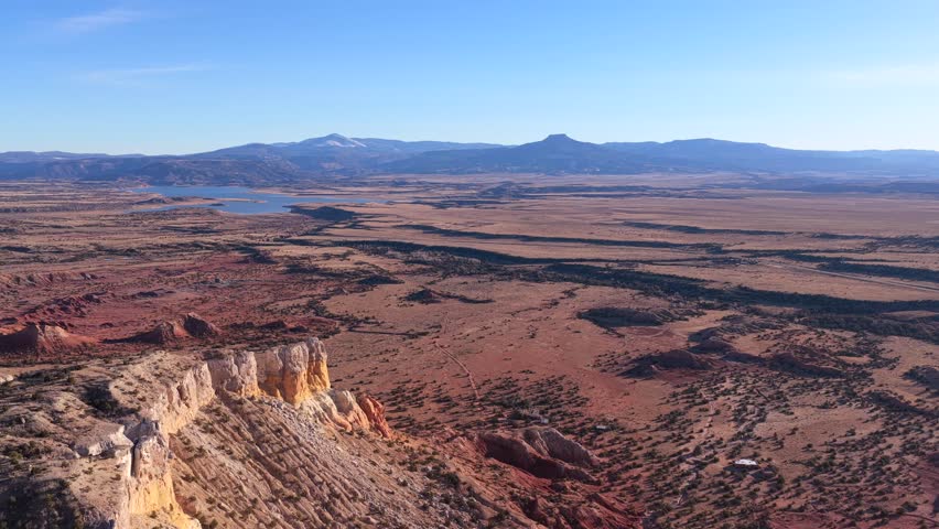 Drone footage floats above open desert plains and scattered red rock formations, revealing winding natural patterns, sparse vegetation, distant water, and Cerro Pedernal anchoring the horizon.
