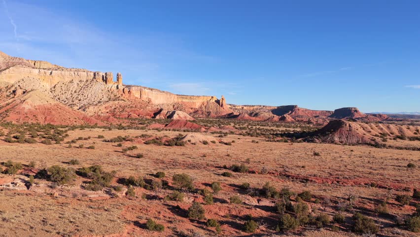 Drone footage glides smoothly over vast arid desert terrain, revealing layered red rock mesas, eroded badlands, sparse vegetation, and expansive open land beneath clear blue skies.