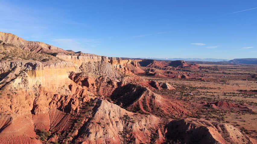 Drone footage moves steadily along eroded red rock cliffs, opening into broad desert badlands with sparse vegetation and repeating geological layers, conveying scale, isolation, and raw natural form.