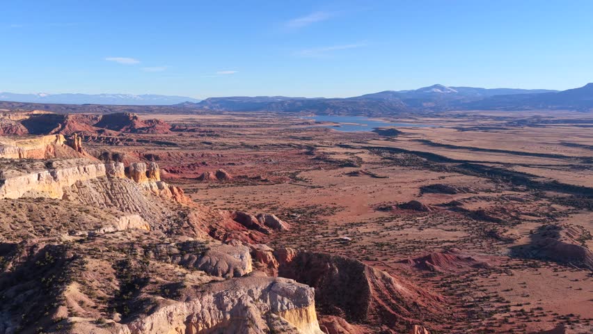 Drone footage drifts above rugged red rock cliffs into sweeping desert plains, revealing layered badlands, scattered shrubs, winding natural patterns, and distant mesas under clear skies.