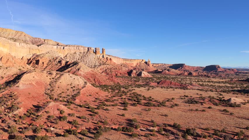 Drone footage glides smoothly above layered red rock mesas and eroded badlands, revealing arid desert plains, sparse vegetation, and dramatic geological forms beneath clear blue skies.