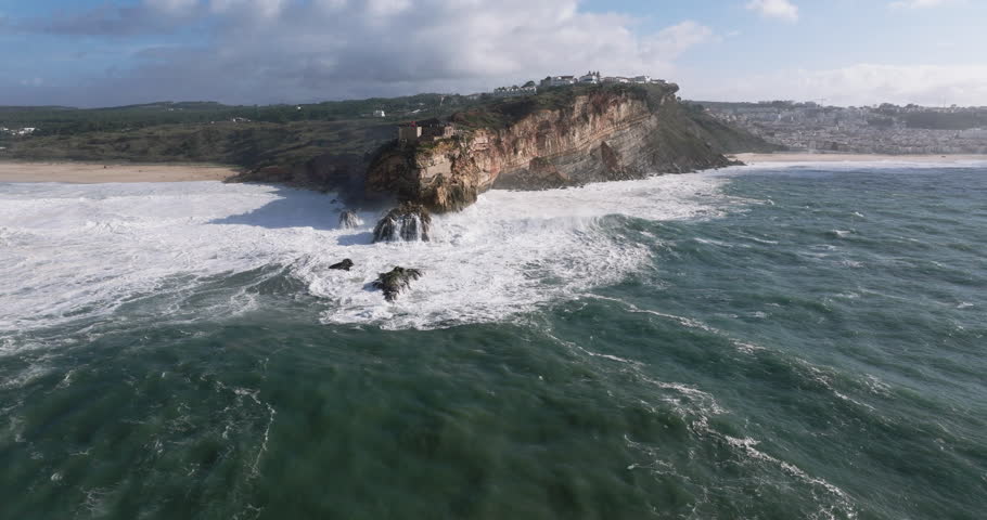 Aerial drone shot of big waves breaking on rocks and cliffs on a day with giant waves in Nazaré, Portugal, Europe. Farol da Nazaré lighthouse and wind turbines visible. Shot in ProRes 422 HQ