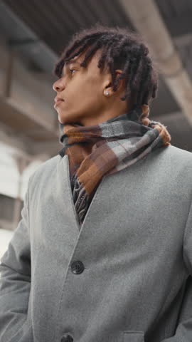 Young black man standing alone on city street and waiting someone, meeting in downtown. Vertical portrait of african american male person in coat looking around, date or gathering with friends