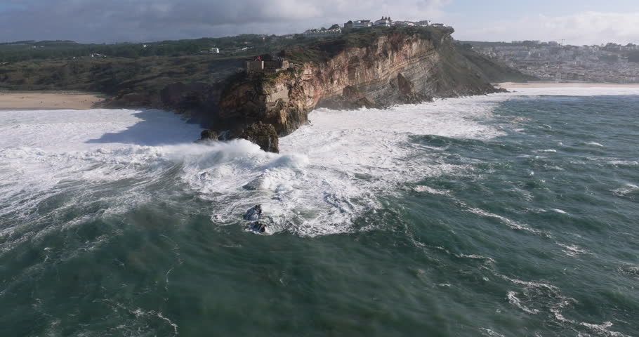 Aerial drone shot of big waves breaking on rocks and cliffs on a day with giant waves in Nazaré, Portugal, Europe. Farol da Nazaré lighthouse and wind turbines