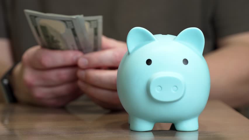Person in the background counting a stack of hundred-dollar bills with a blue piggy bank in the foreground, representing personal finance, investment planning, and wealth accumulation