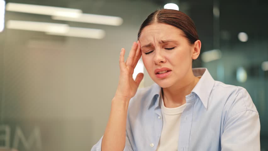 Woman experiences intense headache in office, showing evident pain, discomfort. Feeling stressed, overwhelmed from work pressure, needing relief from throbbing head pain. Health concern, fatigue.