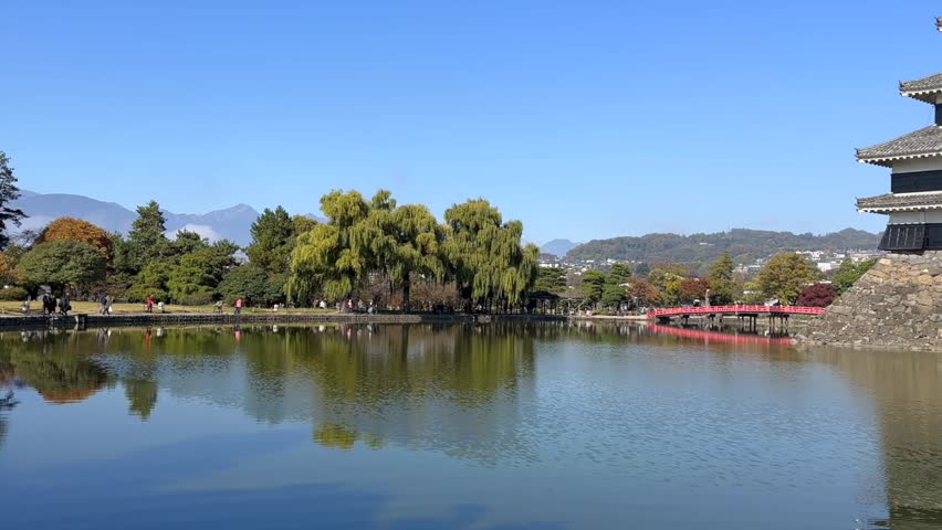 Left to right sweep shows the reflective pond, bright red bridge, and Matsumoto Castle in Japan framed by colorful autumn foliage under a crisp blue sky