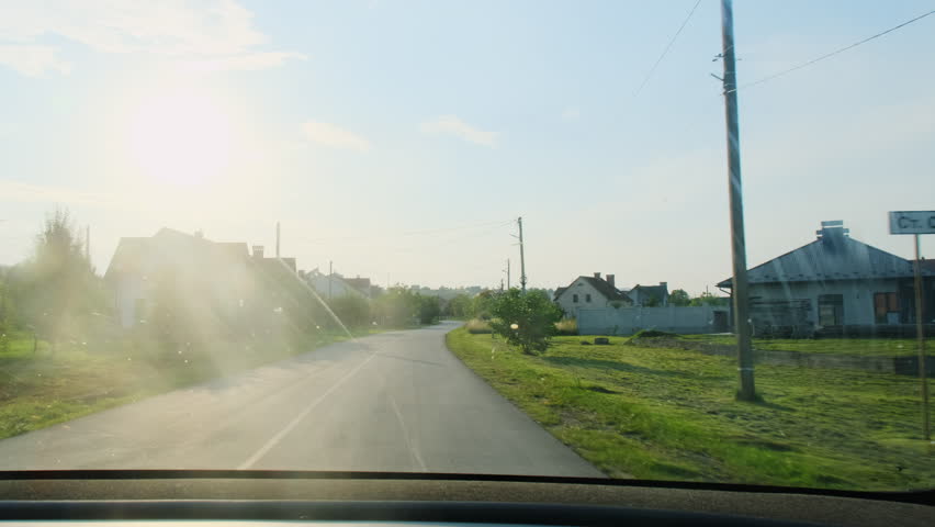 Narrow village road near low old houses through moving car window. Morning sun glares on glass and while neighborhood rolls past slowly
