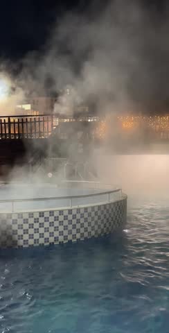 Vertical, nighttime shot of an outdoor thermal pool with heavy steam rising from the hot water into the cold air. Features blue and white tiled pool edge and dim background lighting at a spa resort.