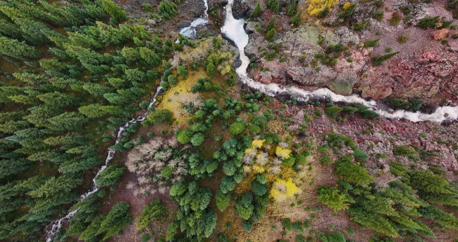 A 4K, top-down drone shot tracking forward over a rushing mountain waterfall in Colorado during autumn, with lush green foliage on one side and vibrant yellow and orange fall colors on the other.