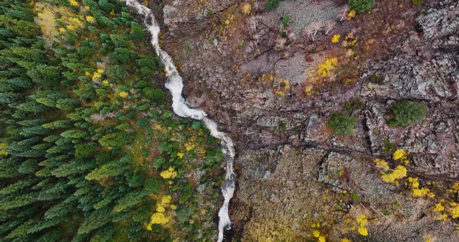 Top-down drone shot tracking forward over a rushing mountain waterfall in Colorado during autumn, with lush green foliage on one side and vibrant yellow and orange fall colors on the other.