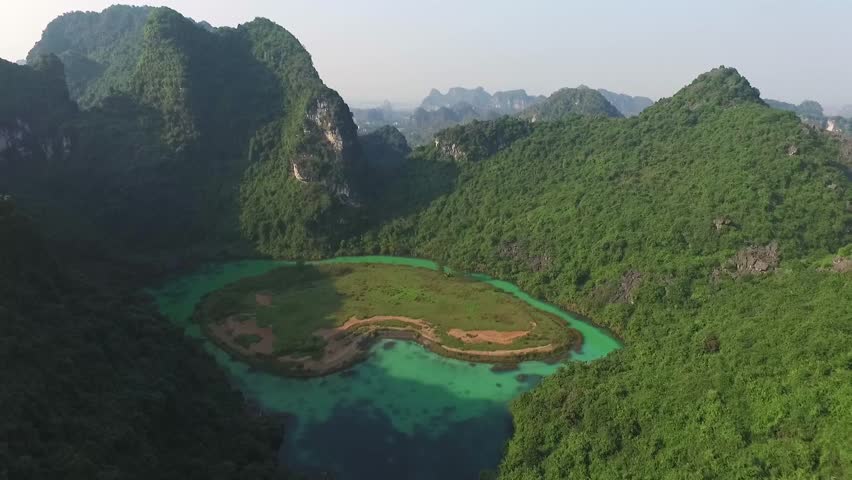 Aerial view of Trang An Valley in Vietnam: A vibrant turquoise lake is nestled between majestic, lushly forested limestone cliffs under a clear, sunny sky, showcasing iconic karst scenery.