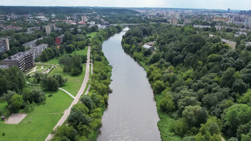 Aerial summer shot of the Neris River in Vilnius, showing green riverbanks, residential districts and walking paths stretching toward the city skyline.