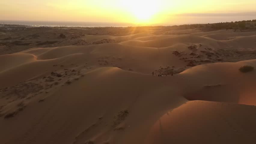 Aerial view of the red sand dunes in Mui Ne at sunset: The low sun casts long shadows over the undulating desert landscape, emphasizing the warm, reddish hues of the dunes.