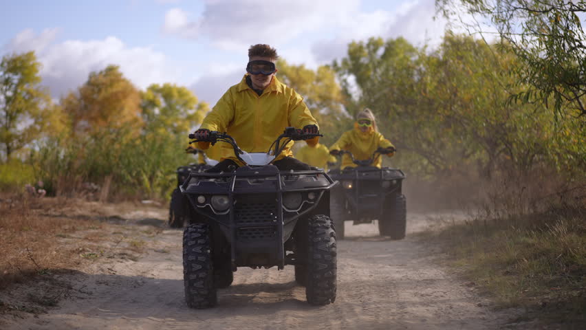 Three riders on ATV trail kicking dust and leaves, yellow jackets and goggles, leader navigating narrow dirt road while partner follows, sunlit autumn trees