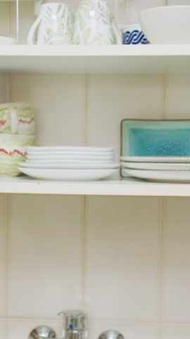 Elderly woman carefully organizing kitchen cupboard, gently placing plates on wooden shelves and softly closing cabinet door during routine household task