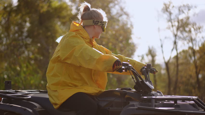 Side profile white rider turning quad through sunlit trees on rural path, adjusting course with calm concentration in yellow jacket and goggles, scenic nature
