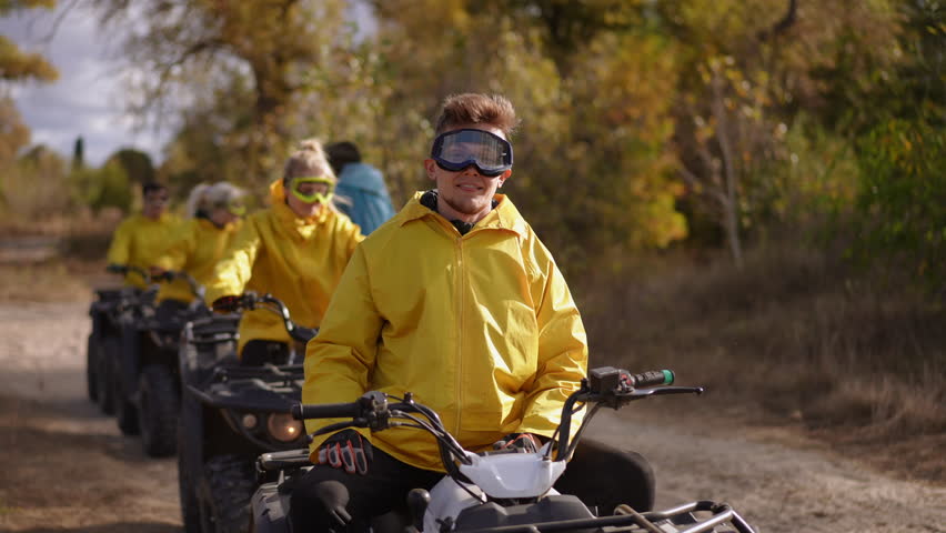 Slow motion. Group of white riders celebrating on quad trail, joyful leader pumps fists while friends in yellow jackets follow in line, dusty sunlit path and upbeat team