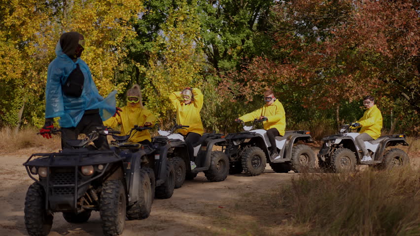 Slow motion. Guided ATV briefing at forest edge leader in blue jacket addresses lineup of riders in yellow jackets beside parked quads crisp autumn foliage calm