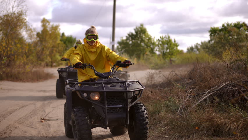 Yellow jacketed riders on atv trail leading convoy, organized patrol along dusty rural road, trees and overcast sky, instructor steering front, two riders