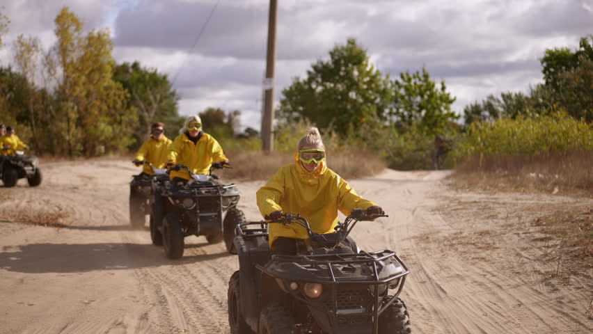 Yellow jacketed riders kicking up dust and gravel on ATV track, lead rider accelerates toward camera, dynamic motion and flying debris, cloudy sky, team chasing