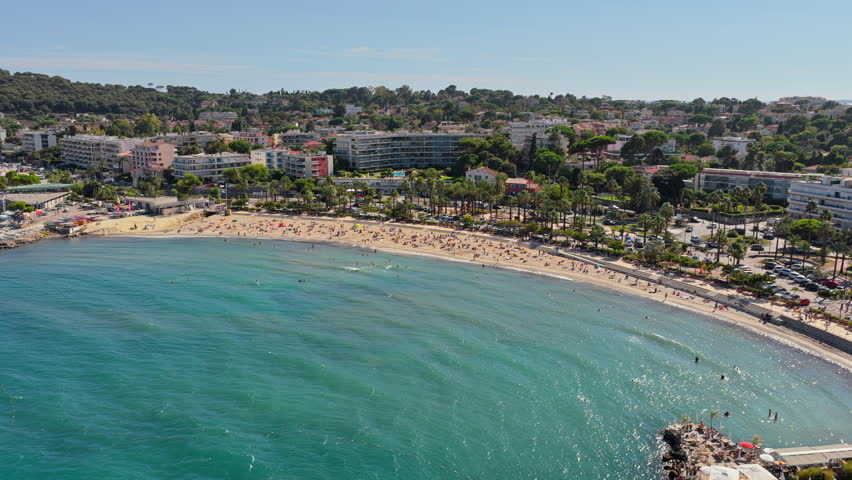 Aerial drone view of Antibes beach and promenade, lined with palm trees and apartments, with turquoise Mediterranean waters and swimmers in the bay