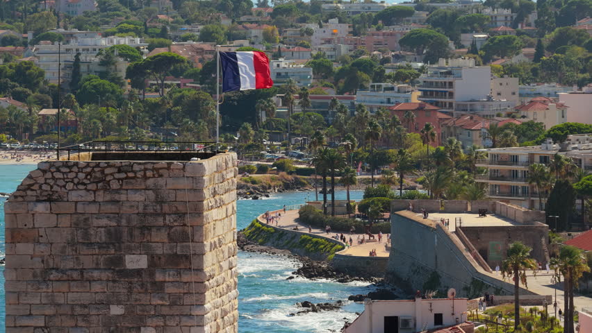 Aerial drone view of the French flag waving above Antibes Old Town medieval tower, with palm-lined promenade and the Mediterranean Sea in the background