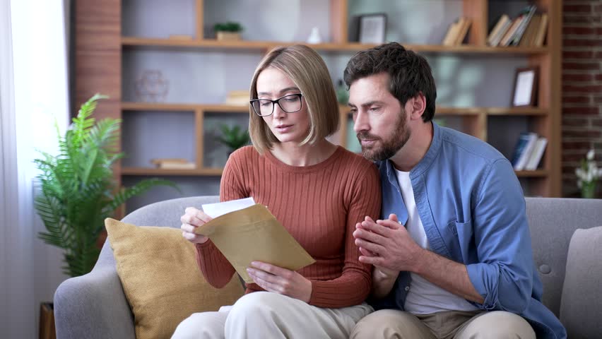 Disappointed married couple reading letter with bad news sitting on sofa in the living room at home. Upset man and woman are shocked after opening the envelope, depressed by an unpleasant notification