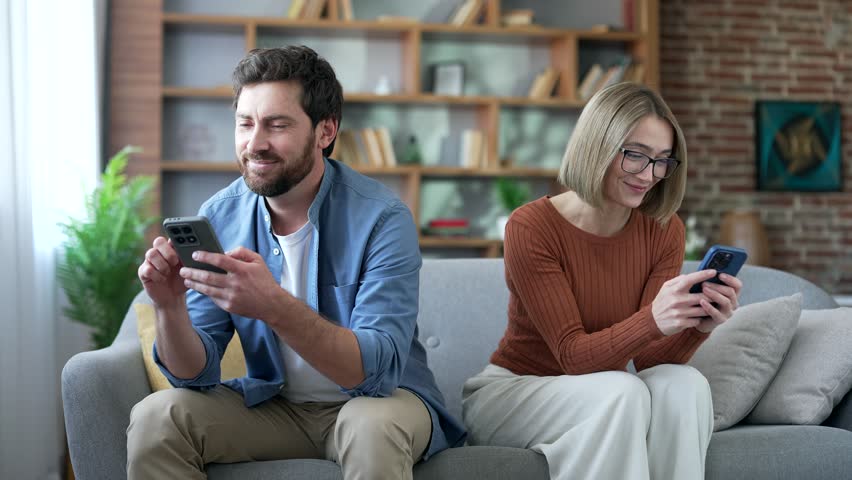 Mobile phone addicted couple sits back-to-back on sofa at home use smartphones. Bored distant family ignoring each other. Relationship problems, overuse addiction to gadgets, internet and social media