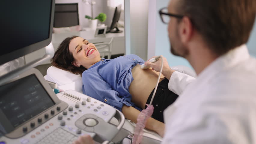 A male doctor in a white coat operates an ultrasound machine while a woman lies on an examination table, looking up at the screen