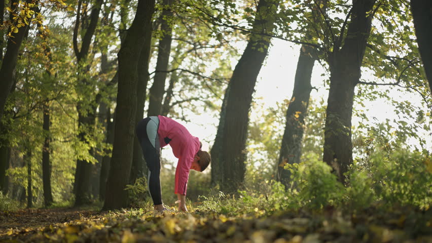 A woman in athletic wear performs a standing forward bend exercise amidst the trees and golden foliage of an autumnal woodland setting