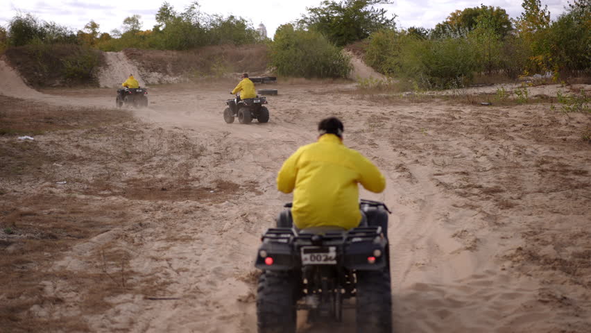 Yellow jacket rider follows dust trail, pack of quads tearing through open sandplain with aggressive throttle, dynamic rear perspective, energetic chase vibe