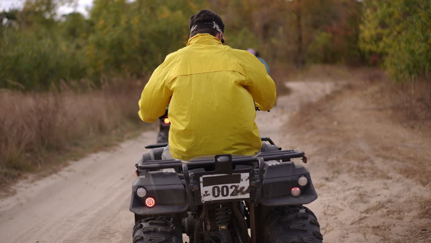 rear view rider yellow jacket atv trail kicking up dust along rural dirt path through autumn forest, helmeted adult navigating winding track with friends ahead,