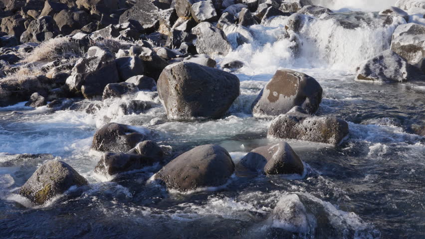 Frozen river with powerful white water rapids flowing over dark, icy rocks and frosted grass in a rugged natural landscape. Winter scenery, cold environment.