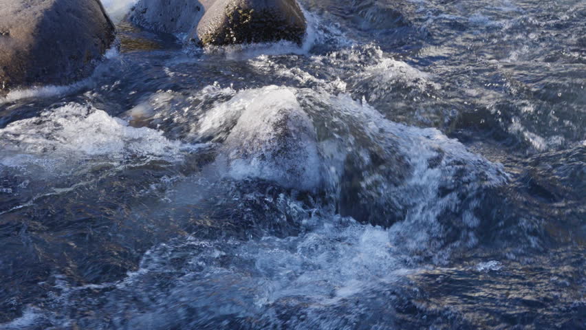 Frozen river with powerful white water rapids flowing over dark, icy rocks and frosted grass in a rugged natural landscape. Winter scenery, cold environment.