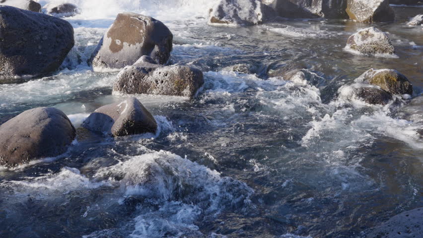 Frozen river with powerful white water rapids flowing over dark, icy rocks and frosted grass in a rugged natural landscape. Winter scenery, cold environment.