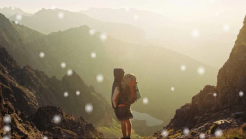 "Hiker with a backpack standing on a rocky mountain peak at sunset, overlooking a vast, misty landscape with a magical snowy bokeh overlay."