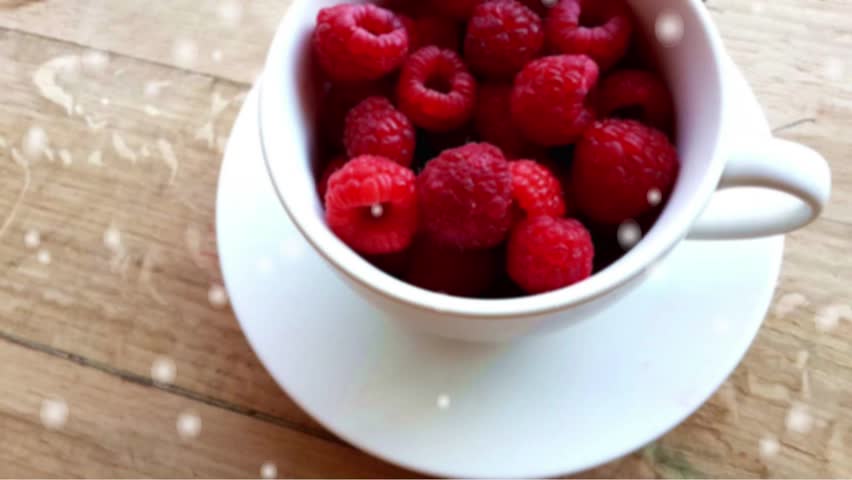 "White cup filled with fresh red raspberries sitting on a wooden table with a soft, blurred white bokeh overlay."