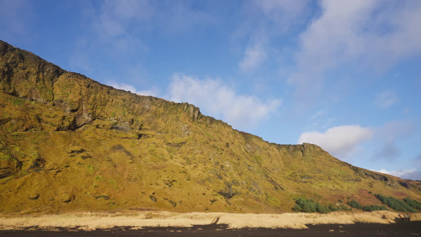 Vast Icelandic landscape featuring a rugged, moss-covered mountain under a dynamic blue sky with moving clouds. Golden grass and black sand foreground hint at a volcanic region.