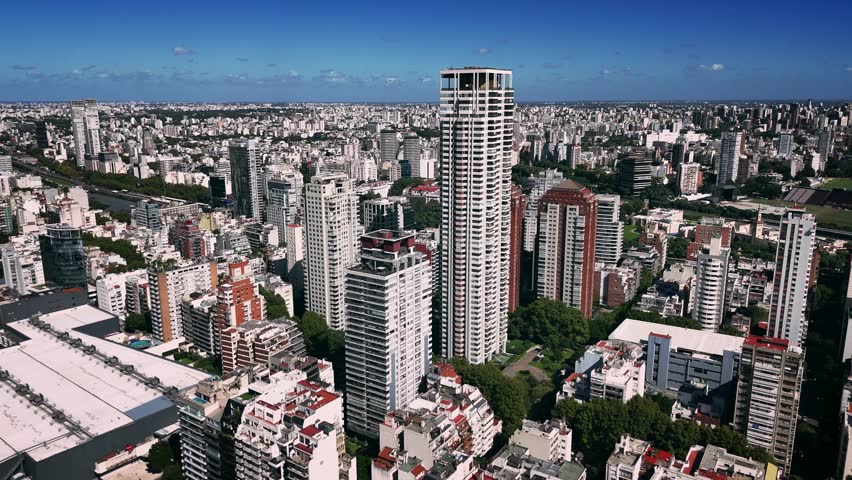Aerial drone view of Buenos Aires, Argentina, featuring a prominent modern residential skyscraper rising above a dense urban landscape. The image showcases the city’s vertical growth, contemporary architecture, and surrounding neighborhoods with tree-lined streets and mixed-use buildings. This high-altitude perspective highlights metropolitan expansion, real estate development, and the dynamic character of one of South America’s largest capital cities.