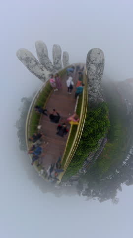 Vertical video. Golden Bridge Forming Tiny Planet Effect in Dense Fog with Giant Stone Hands Holding Curved Walkway on Bana Hills during Surreal Landmark Scene.