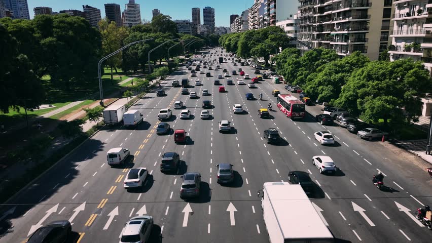 This aerial image shows a wide avenue in Buenos Aires, Argentina, captured from an elevated perspective that reveals the scale and structure of everyday life in a major South American capital. The multi-lane roadway is filled with moving vehicles, creating a strong sense of motion, order, and connectivity during daytime.

On one side of the avenue, a continuous line of residential buildings rises along the street, featuring balconies, repetitive windows, and a functional contemporary design. These structures reflect high population density and residential development, framing the avenue as a central axis for movement and daily activity.

On the opposite side, a large green area with mature trees provides visual contrast and balance. This natural space highlights the relationship between infrastructure and public green areas, showing how transportation routes coexist with recreational and environmental zones. The contrast between asphalt, buildings, and vegetation adds depth and visual harmony to the scene.

Natural daylight enhances clarity, sharpness, and color accuracy, making the image clean and realistic. The elevated viewpoint allows a clear understanding of traffic flow, road layout, and surrounding architecture, offering a comprehensive perspective that cannot be experienced from street level.

This image communicates themes such as transportation systems, mobility, daily commute, infrastructure, and modern lifestyle. It is suitable for editorial or commercial use related to travel, business, development, or mobility concepts. Overall, the photograph conveys movement, structure, and organization, capturing the character of Buenos Aires from above in a strong and versatile composition.