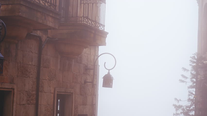 Foggy alley between stone buildings with lanterns hanging above railing and soft pale sky creating quiet atmospheric European style street mood.